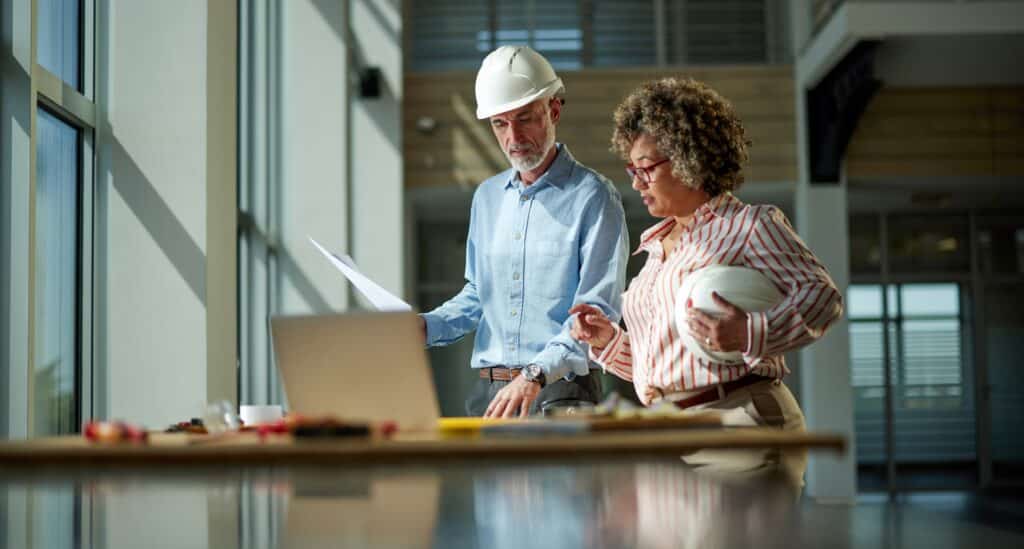 A man and a woman colleague at a construction site discuss building plans in a brightly lit space.