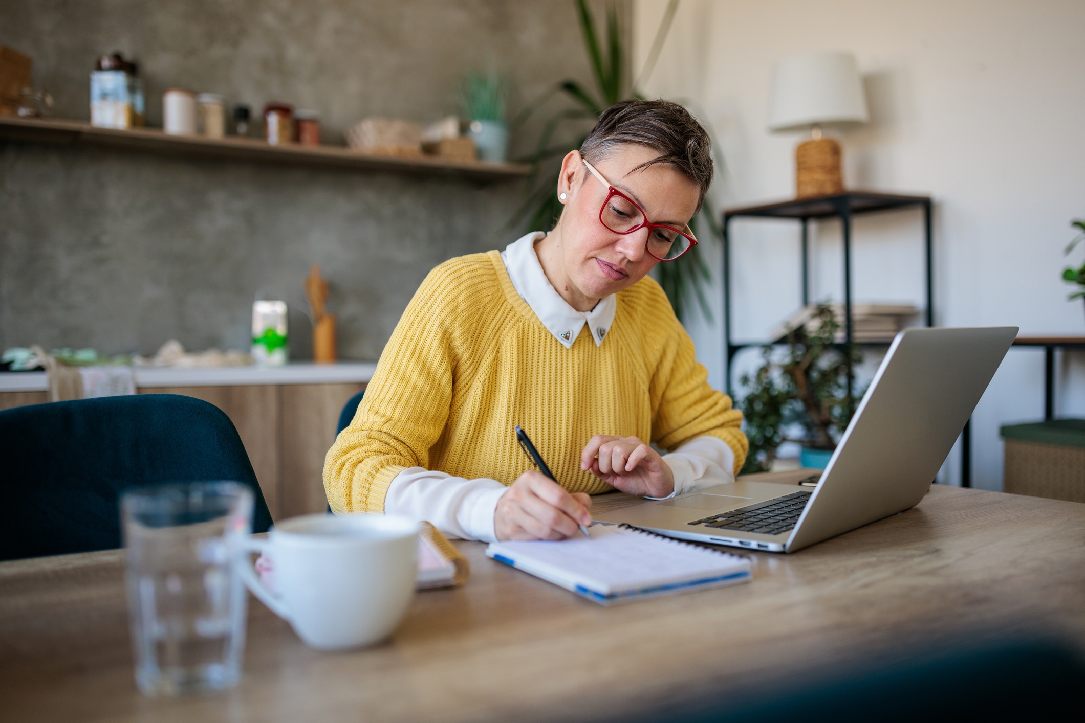 Person sitting at a table, with a laptop open in front of them, and taking notes in a notepad 