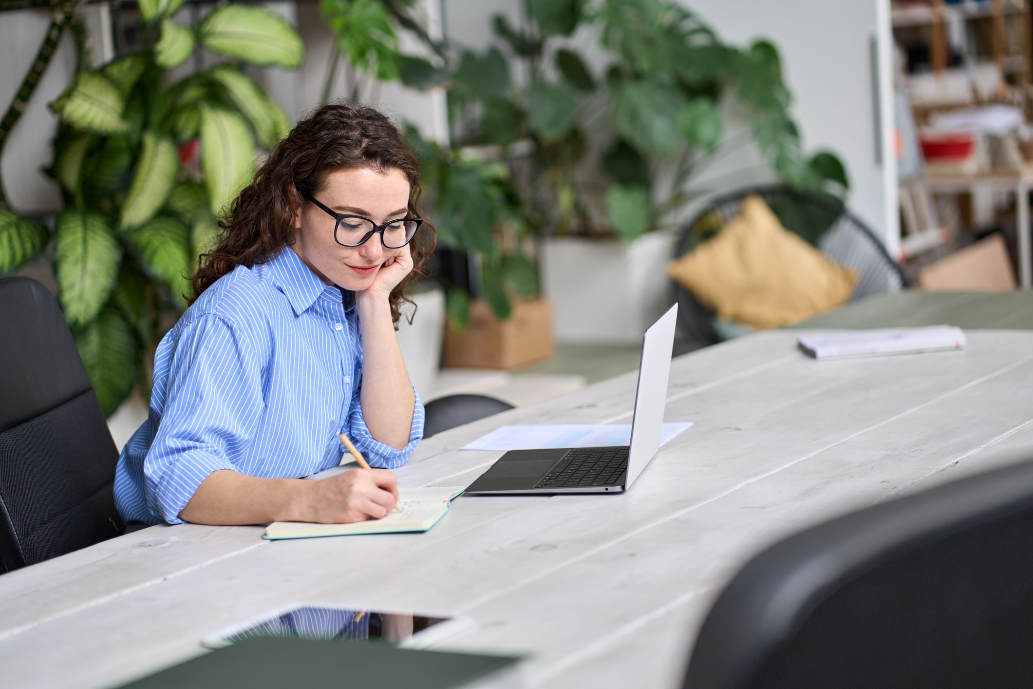 Young professional or student smiling and writing in a notebook next to an open laptop.