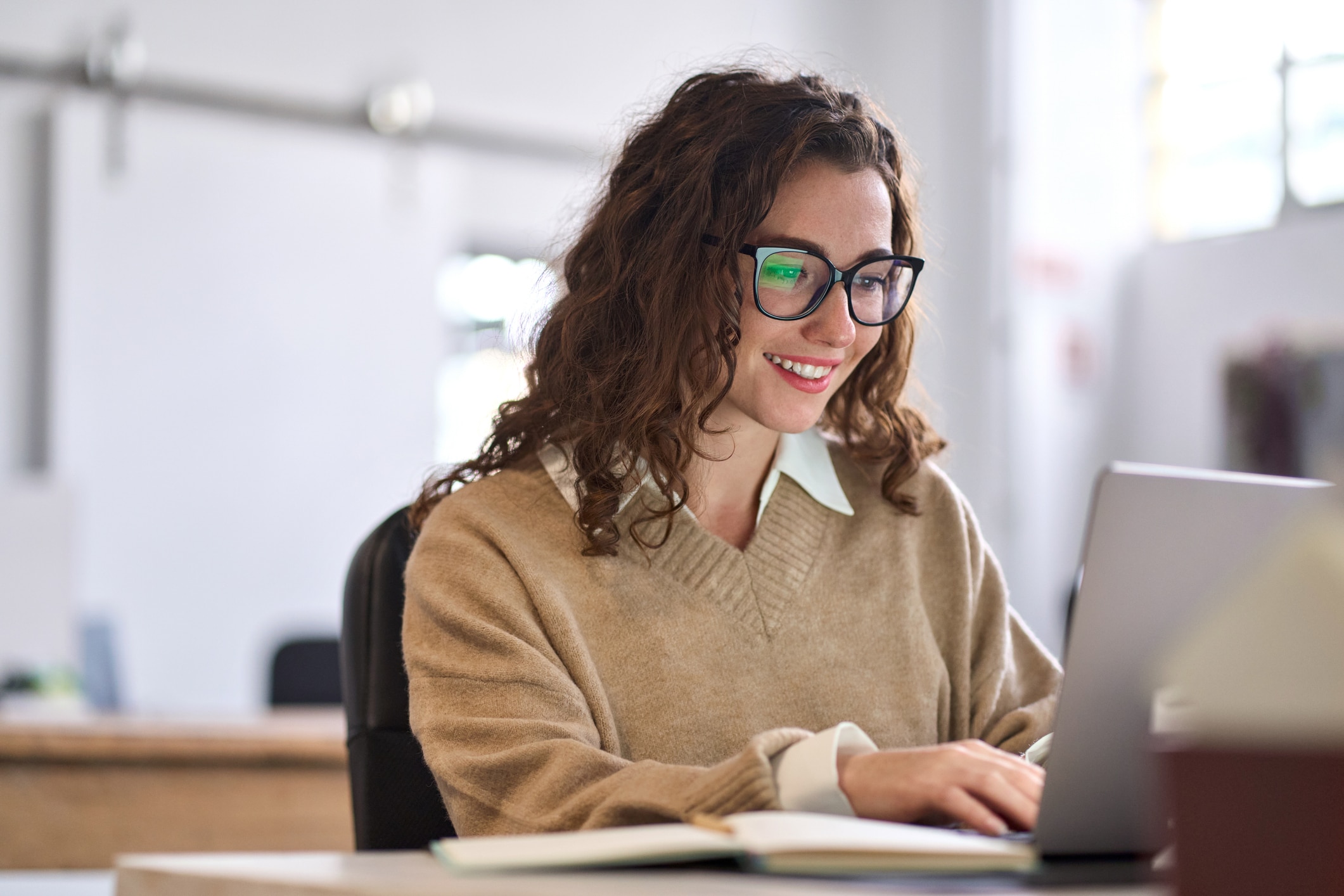 Young happy professional sitting at desk working on laptop in corporate office