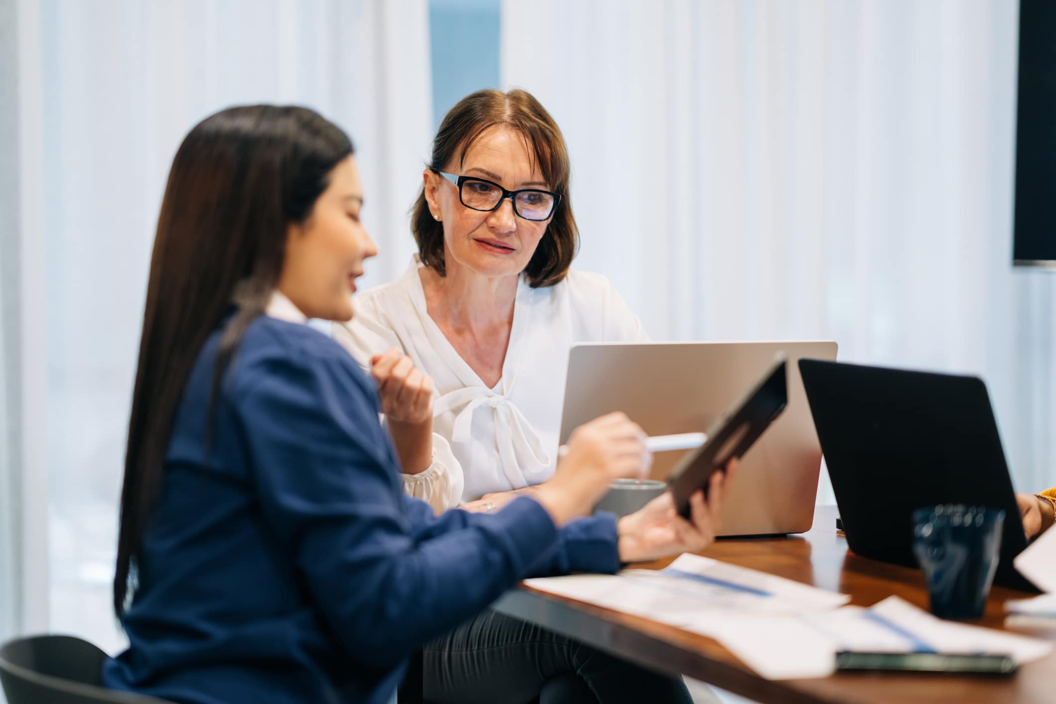 Two professionals sitting at a wooden table in a well-lit office space, engaged in a focused work conversation. One person, holding a digital tablet, explains or presents something to their colleague, who listens attentively while seated in front of an open laptop. Documents, coffee mugs, and a second laptop are visible on the table, suggesting an ongoing collaborative work session. The setting feels modern and relaxed, with sheer curtains allowing natural daylight to soften the professional environment. 