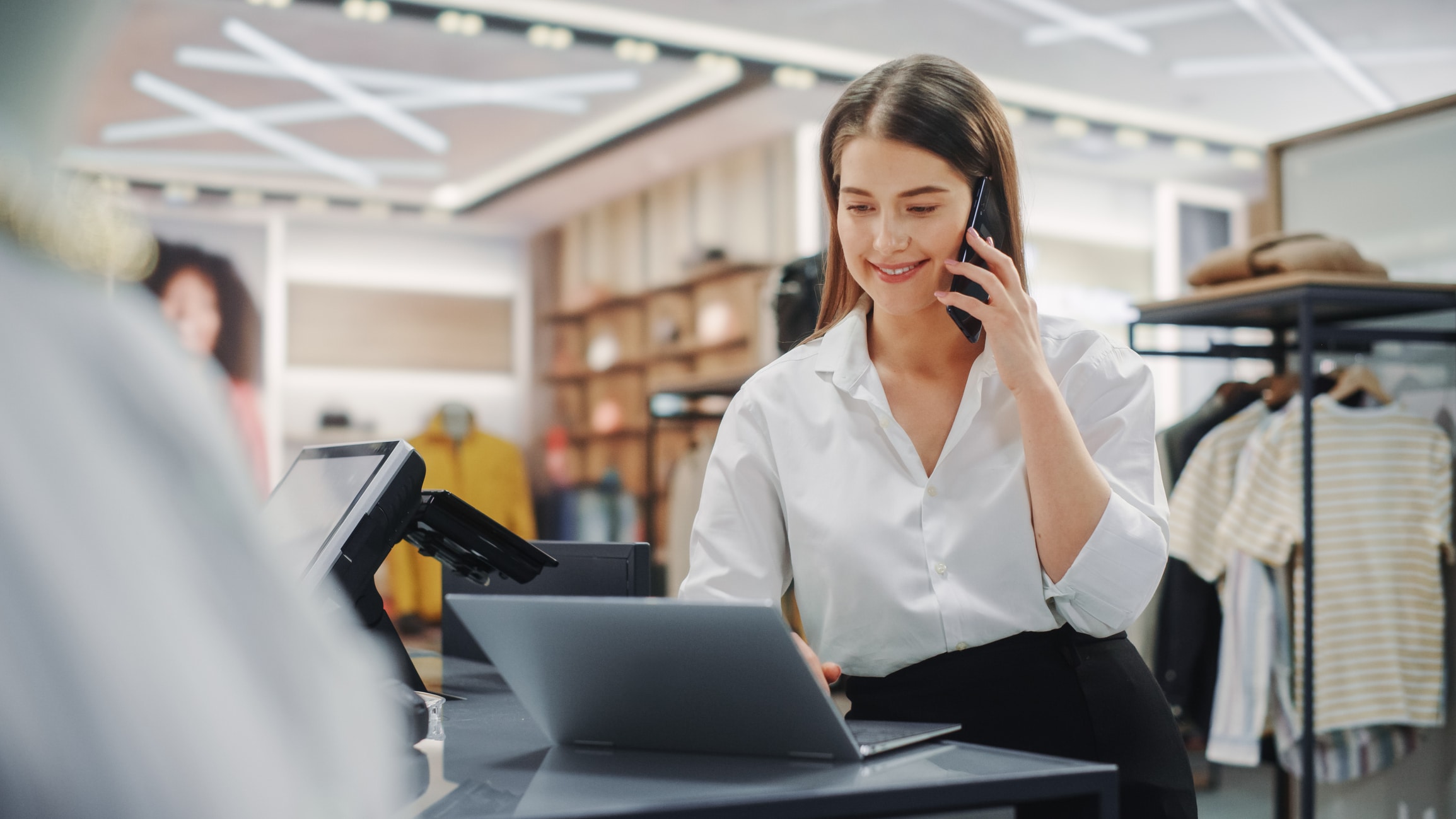 Smiling person working on a laptop and talking on the phone at the same time in a store setting