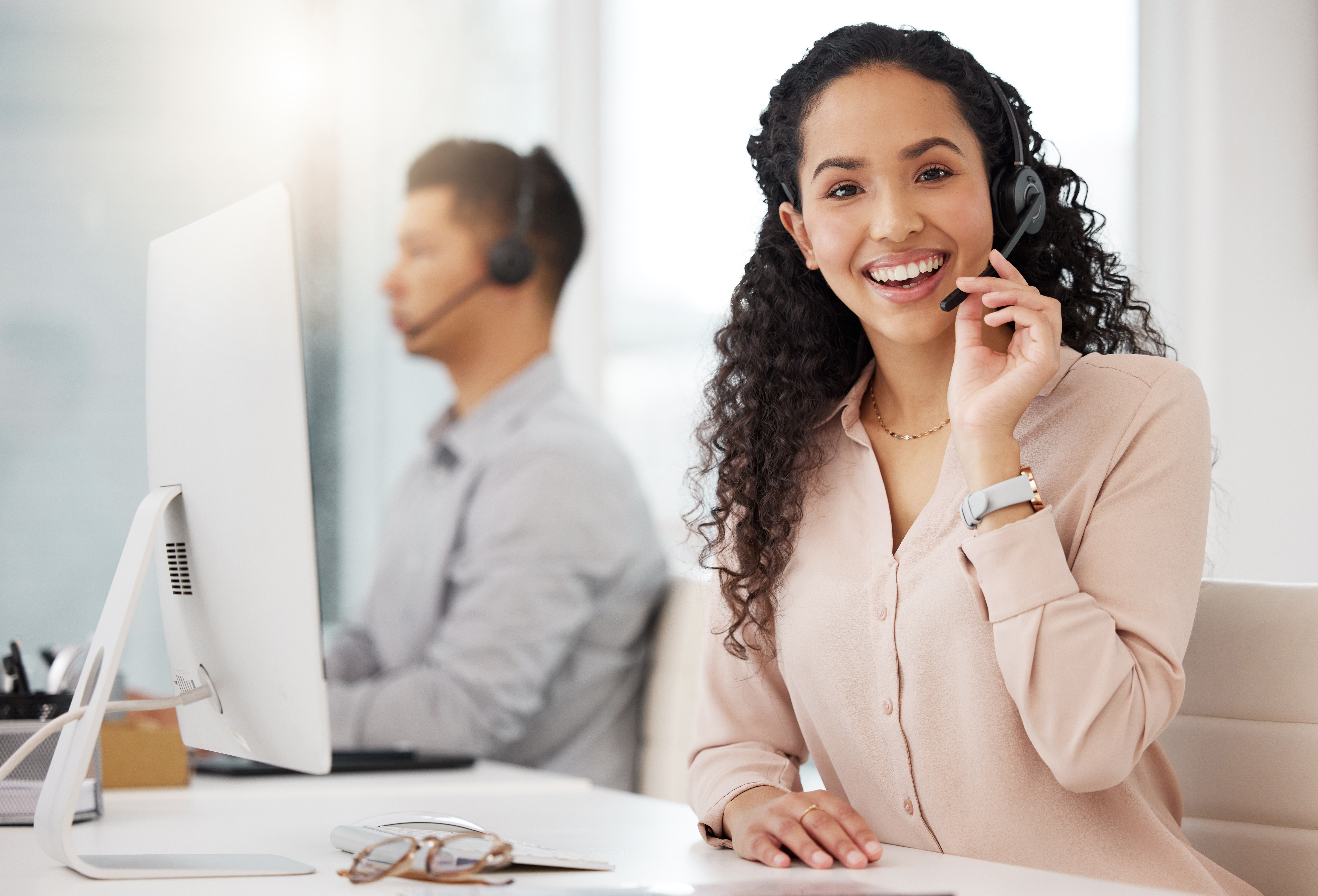 A smiling woman wearing a headset works at a customer service desk, with a colleague visible in the background.