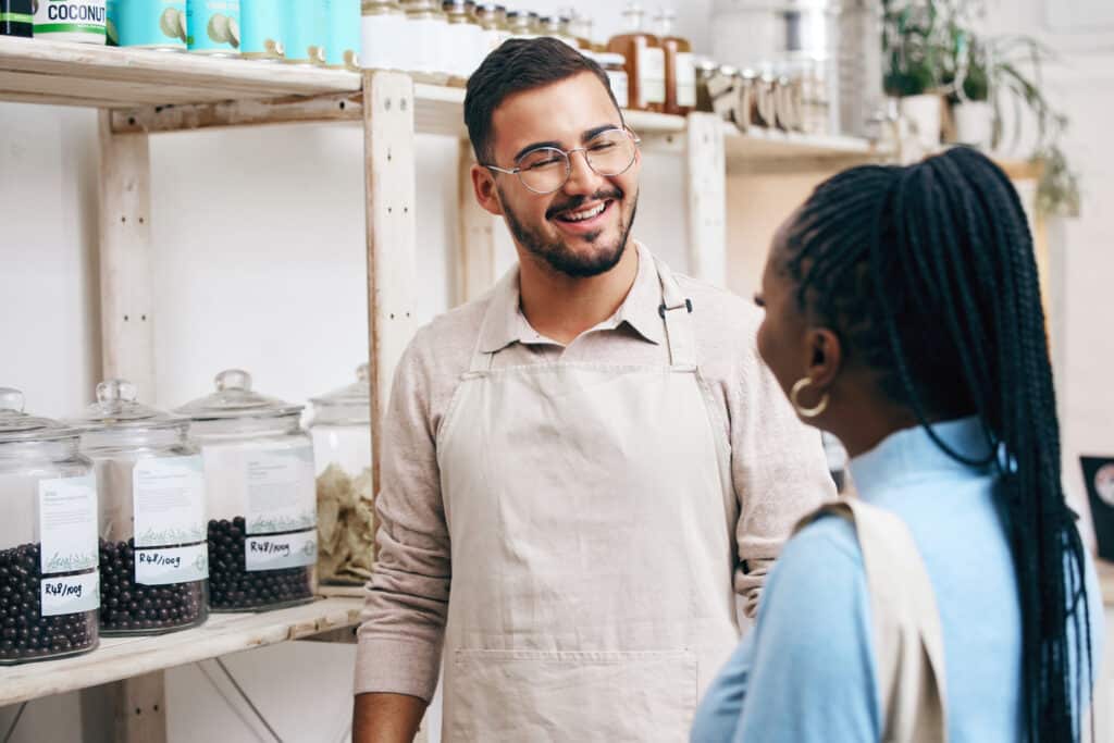 A smiling shop employee in an apron chats with a customer in a specialty grocery store.