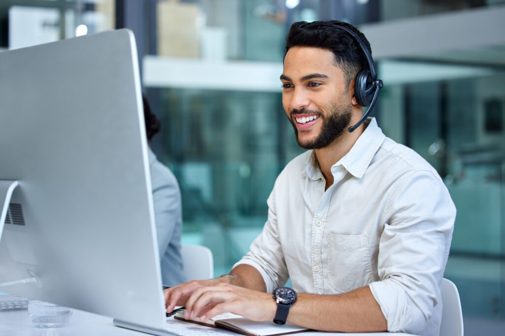 Smiling customer service agent wearing a headset and typing at a desktop computer in an office.