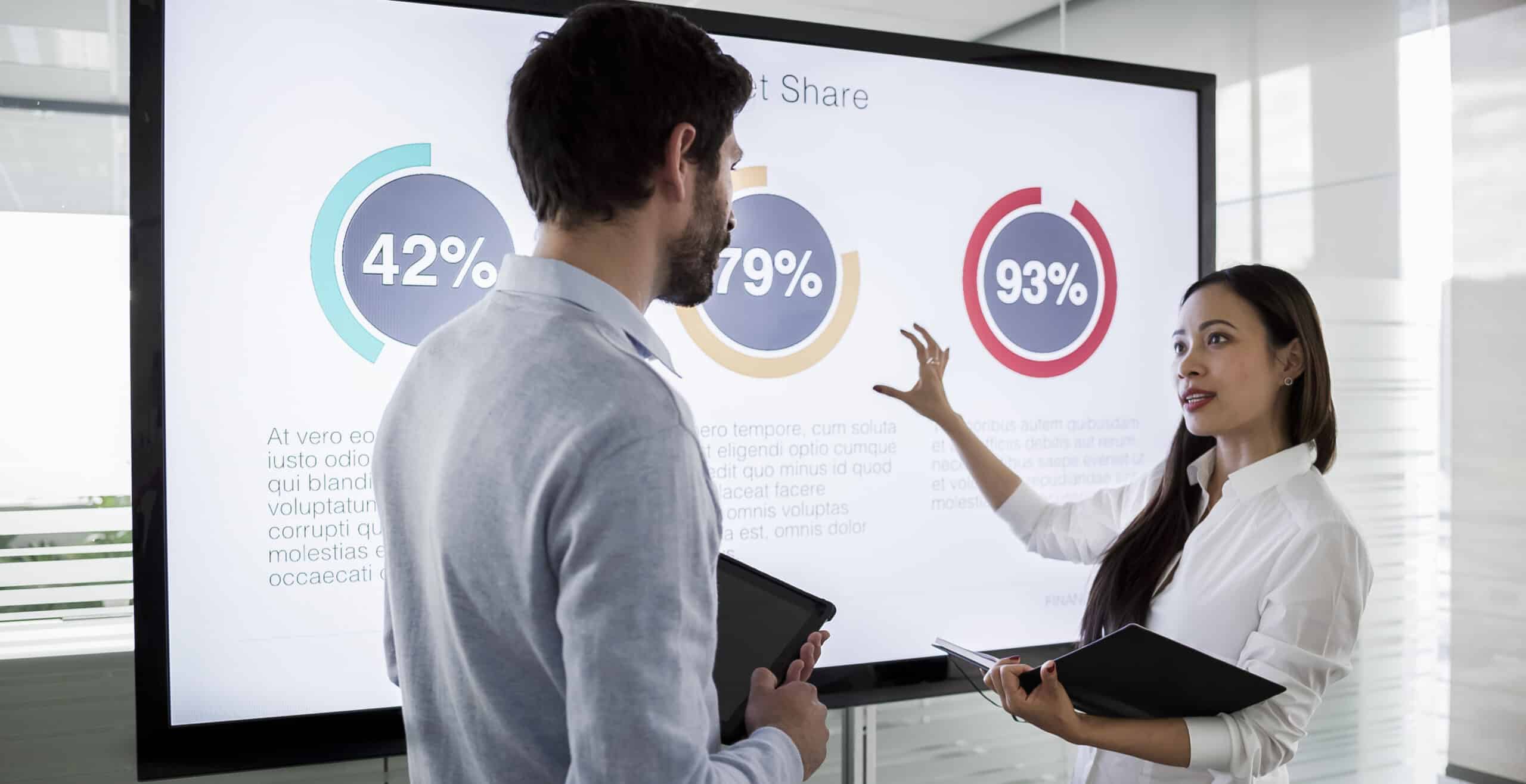 Colleagues standing by large screen in meeting room, going over financial presentation shown on screen in meeting room.